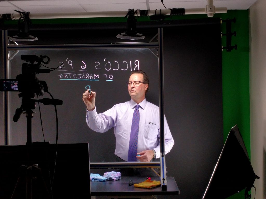 A male professor in a dress shirt and tie is writing on a lightboard with a marker in a video recording studio. The text on the lightboard is reversed due to the camera perspective. The studio is equipped with professional lighting and video recording equipment, and there is a green screen in the background